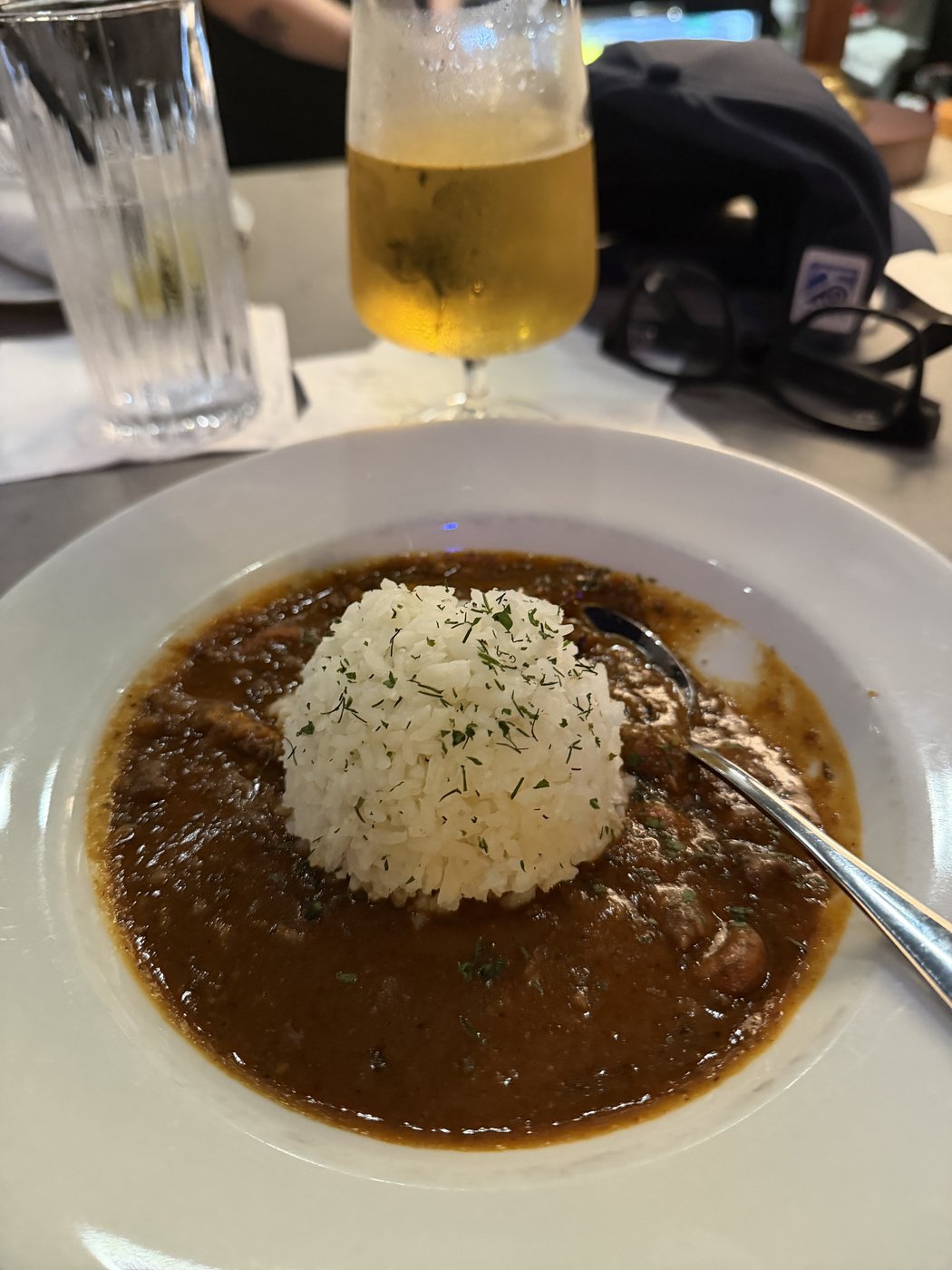 A bowl of sausage and crayfish étouffée with a mound of rice and a glass of beer on the bar