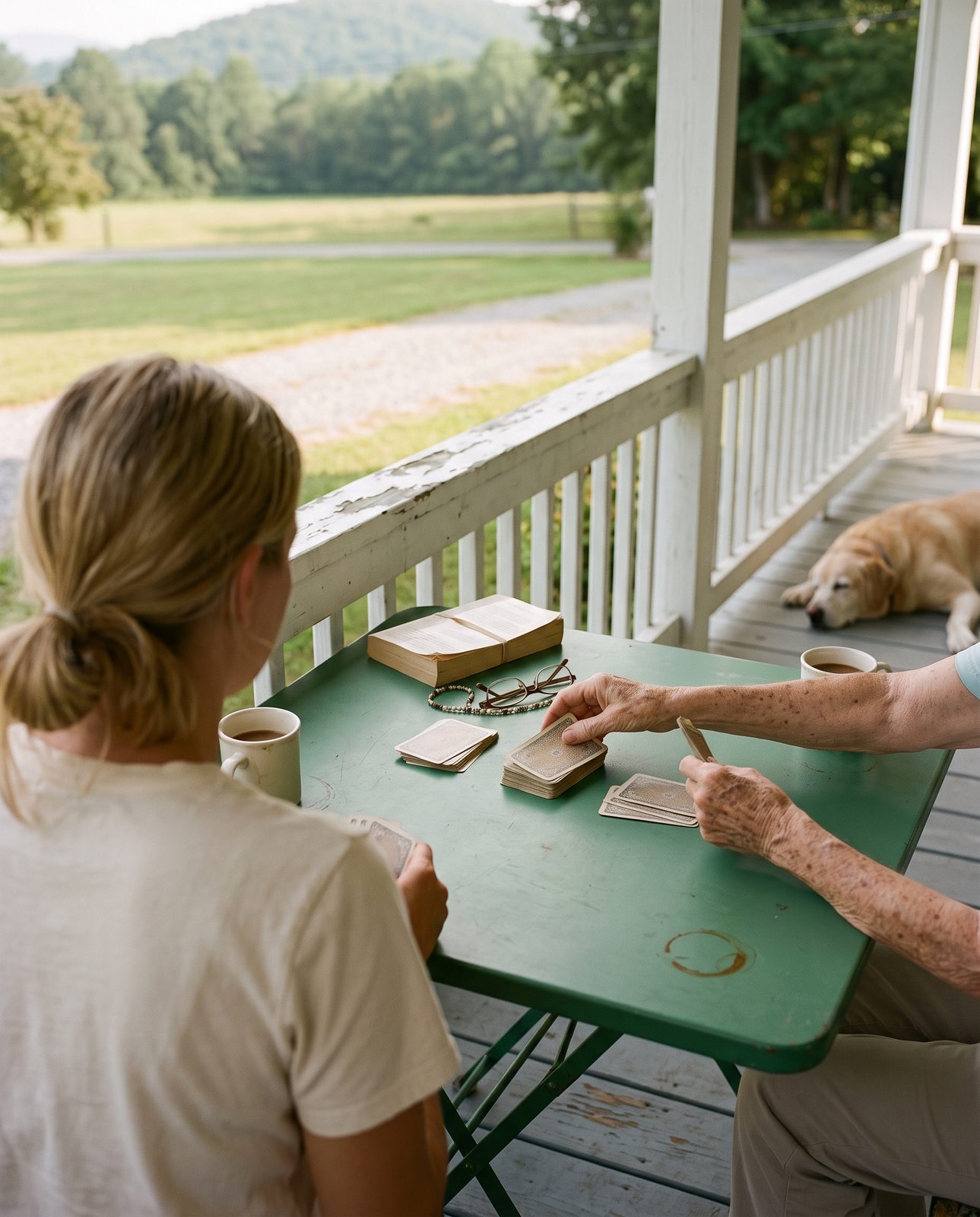 An older woman's hands dealing a worn deck of cards across a green painted metal folding table on a shaded porch, reading glasses on a beaded chain and a creased paperback beside them, a younger woman seen from behind holding her cards low, an old yellow lab soft and cropped at the edge of the boards, mowed field and gravel drive beyond the rail