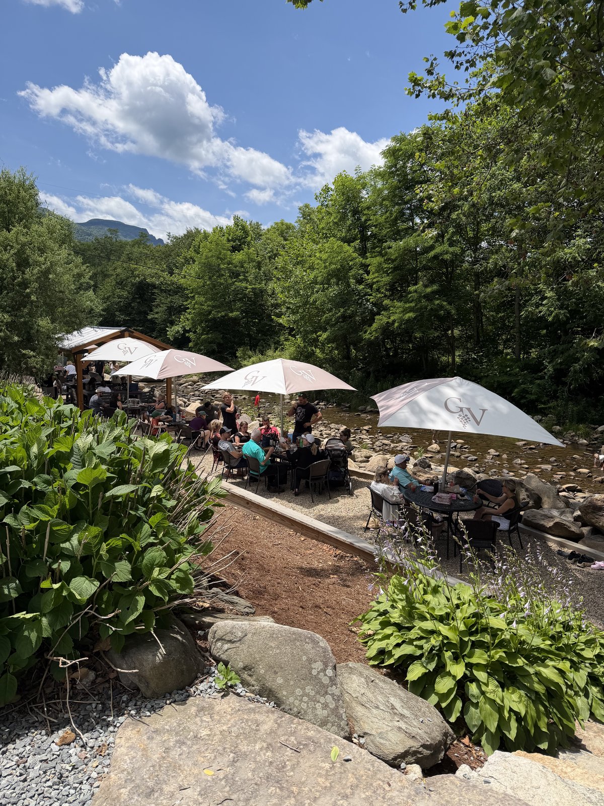 A creekside patio at a mountain vineyard near Banner Elk, NC — umbrellas along the Watauga River with Grandfather Mountain going soft behind under a summer sky