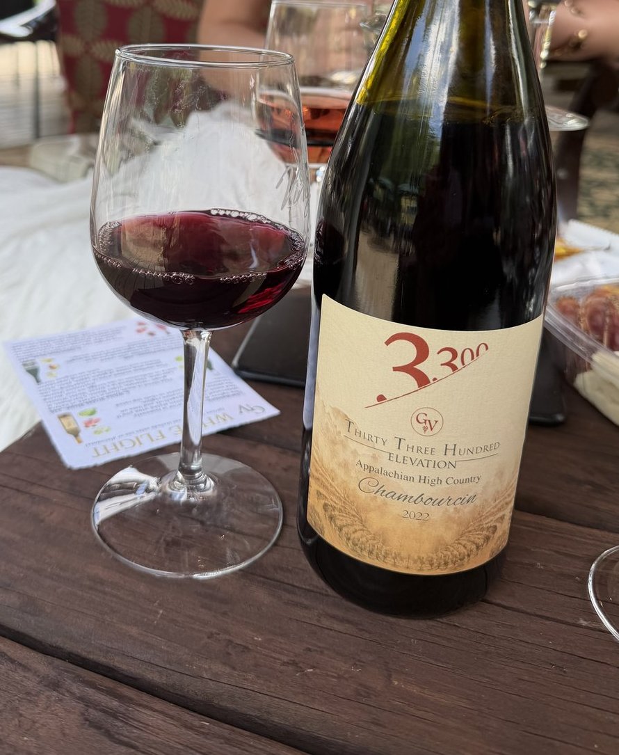 A bottle of Thirty-Three Hundred Chambourcin and a poured glass of red on a weathered crate, the creekside patio behind