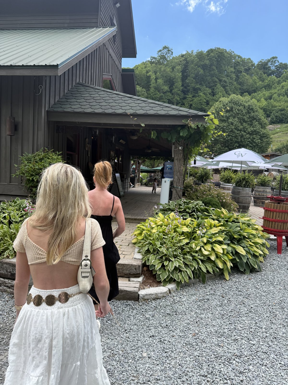 Two figures walking in from behind toward the tasting room, gravel path, hostas and wine barrels alongside, no faces