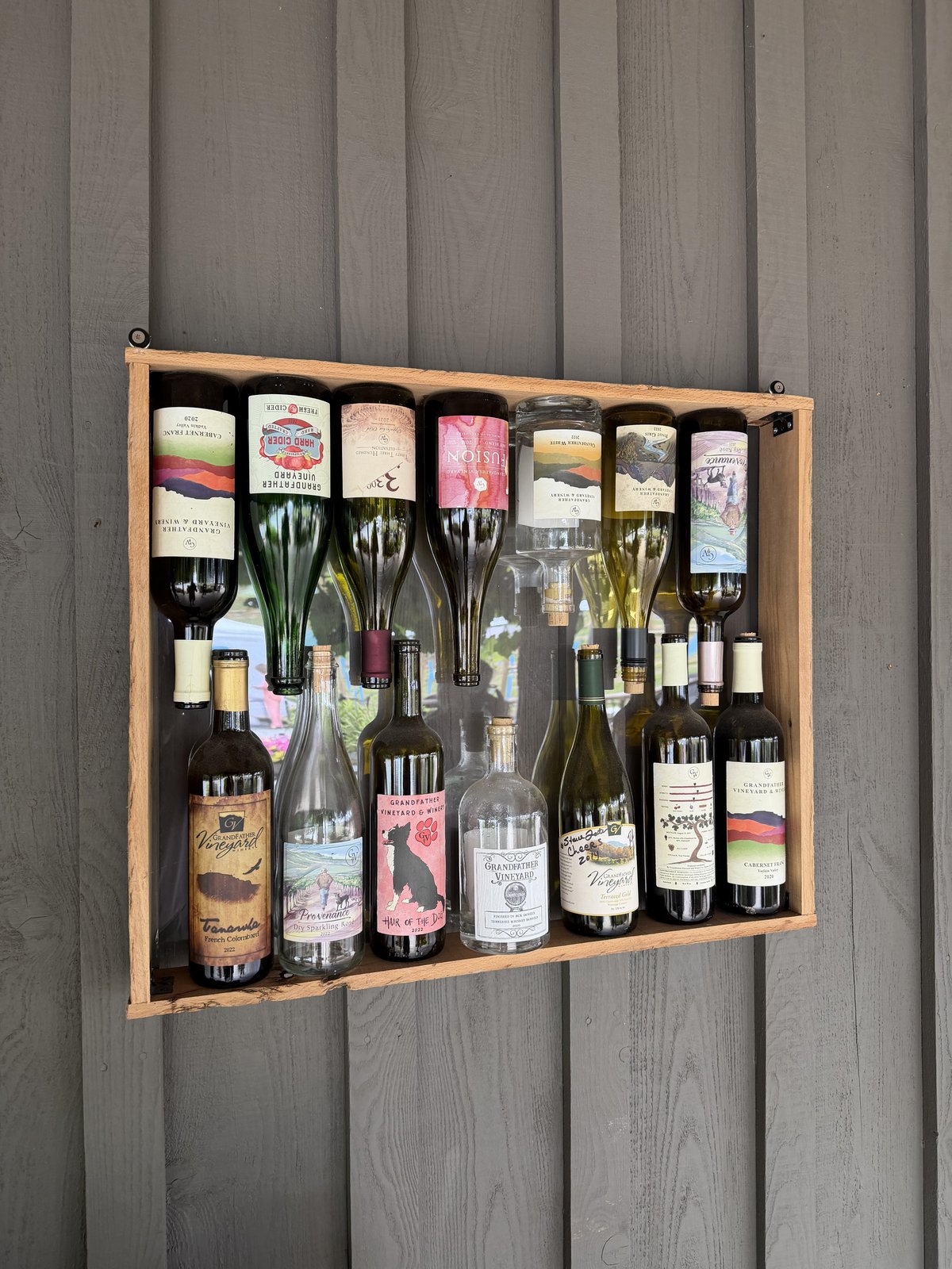 A wall rack displaying the full range of Grandfather Vineyard bottles, empties turned label-out on weathered gray siding