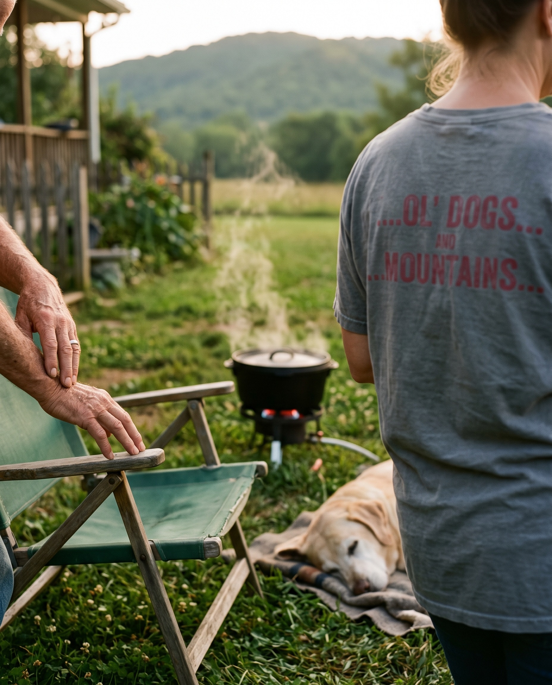 A woman seen from behind in a faded gray t-shirt in a side yard at golden hour, an older man's hands resting on the arm of a green canvas folding chair at the left edge of the frame with no face visible, a cast-iron pot steaming on a low propane burner in the grass, an old yellow lab asleep on a folded blanket, garden fence and porch to the left, a mown path running back to one low tree-covered ridge