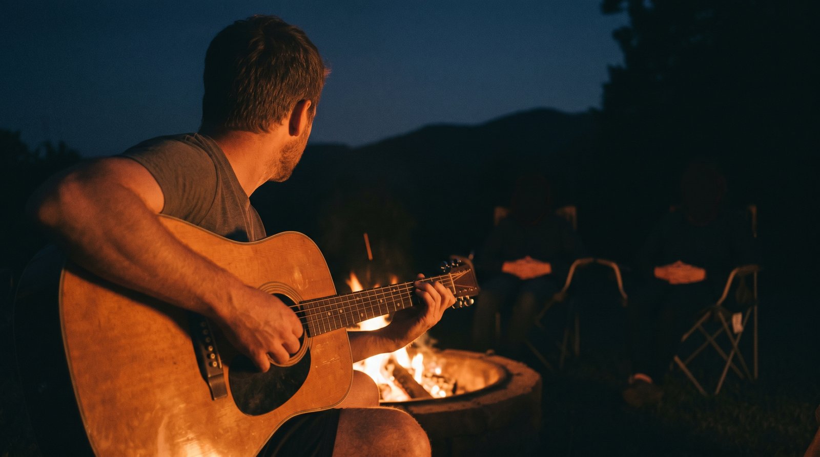 Wes playing guitar at the fire pit after dark, the family around the low flames