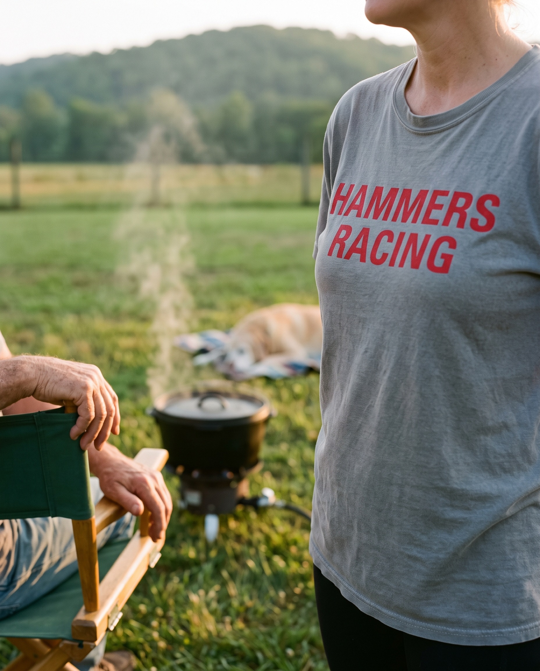A woman in a faded gray t-shirt printed HAMMERS RACING in red block capitals across the chest, standing in a side yard at golden hour with her face out of frame, an older man's hands resting on the arm of a green canvas folding chair, a cast-iron pot steaming on a low propane burner, an old yellow lab asleep on a blanket in the middle distance, one low tree-covered ridge behind
